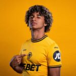 Portrait of a young man in a yellow Wolves jersey against a solid yellow background, pointing to the club crest on his chest.