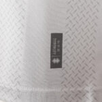Close-up of light gray textured fabric with a small black tag featuring vertical text and a diamond-shaped logo.