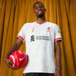 Portrait of a Liverpool FC player in a white away kit, holding a red soccer ball against a mustard curtain backdrop, smiling confidently.