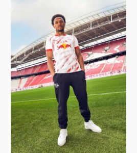 Young man wearing a white Red Bull soccer jersey and navy pants stands on a green stadium field beside red seats.