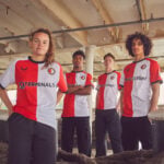 Four young soccer players in red-and-white jerseys pose for a team photo in an unfinished concrete space, hands on hips or crossed arms, looking at the camera.