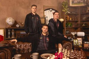 Three men in matching black team jackets pose in a vintage study with a chessboard in the foreground.