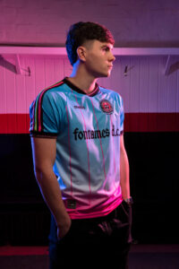 E5277D5A-6FA2-47FC-AA7F-69F7E7D873E7 | UAE SPORT STORE Young man in a light blue football jersey stands in a dimly lit room with pink and blue lighting, hands in pockets, looking to the right