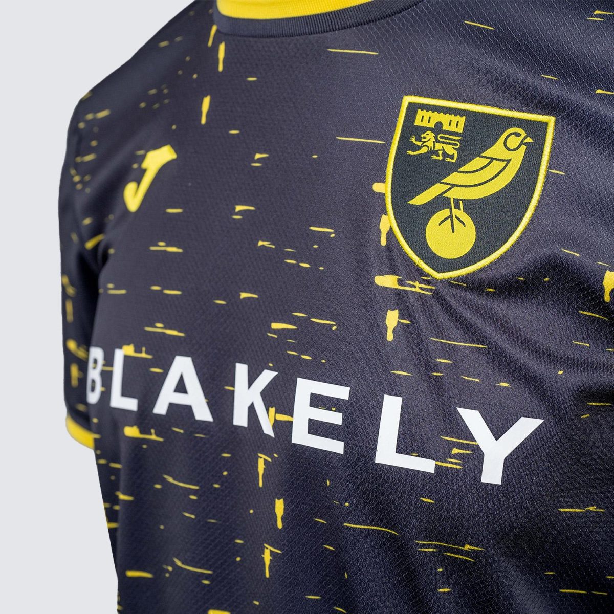Close-up of a dark jersey with yellow speckled pattern, showing a yellow-bordered crest featuring a bird and castle on the chest.