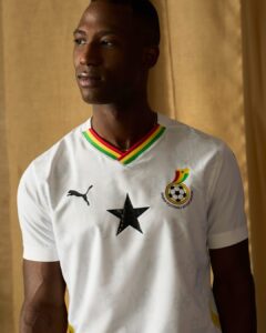 Portrait of a Black man in a white Ghana national football team jersey with star emblem and rainbow collar, looking to the left against a beige curtain.
