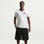 Model wearing a white England football jersey with crest and black shorts, standing with hands in pockets against a light background.