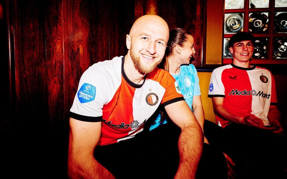 Three teammates sit together, smiling at the camera; left is a bald man in a red‑white jersey, middle a woman in blue, right a man in a red/white jersey.