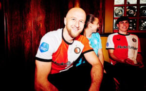 Three teammates sit together, smiling at the camera; left is a bald man in a red‑white jersey, middle a woman in blue, right a man in a red/white jersey.