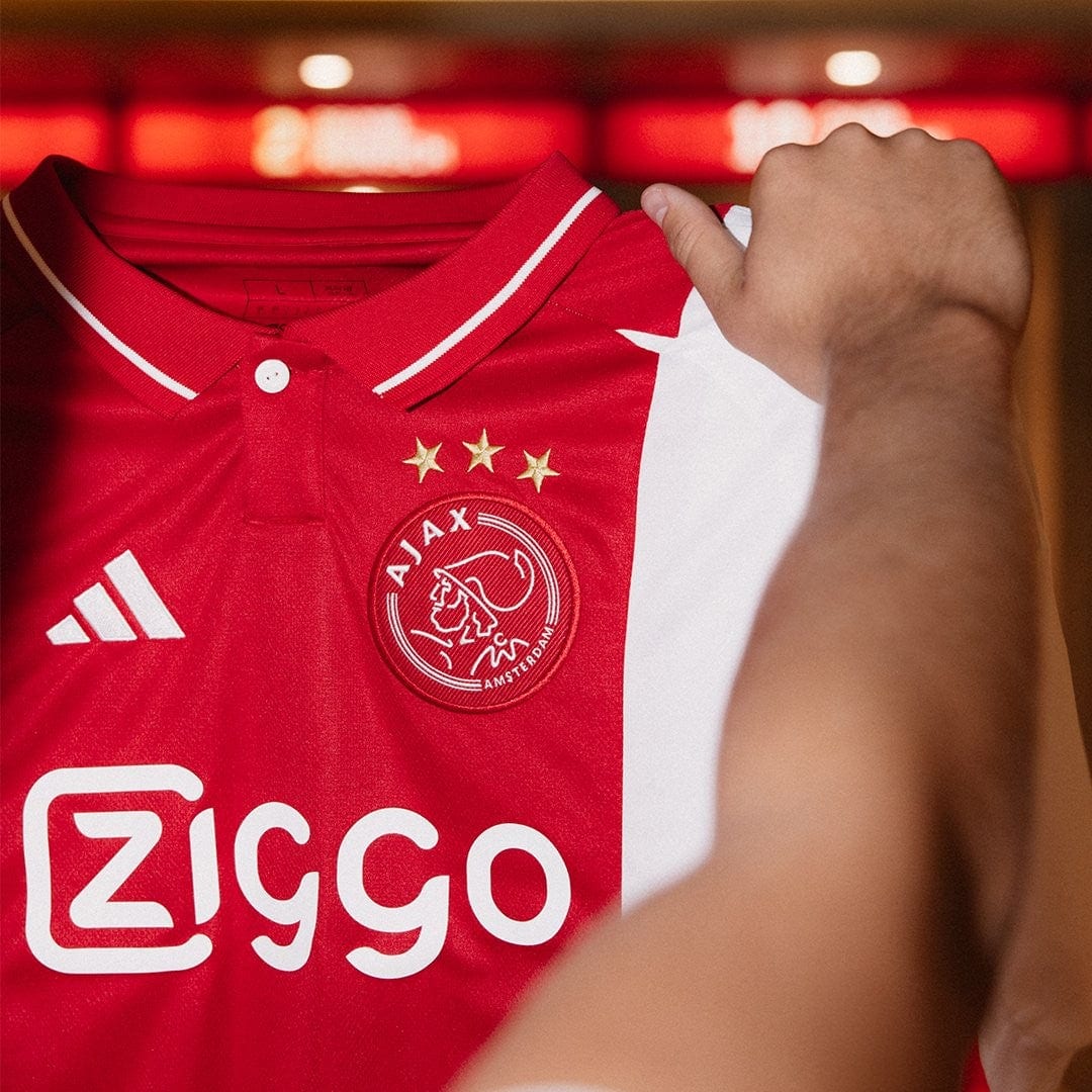 EkOQBqc0vKPuWL4 | UAE SPORT STORE Red Ajax Amsterdam home football jersey being held up, displaying the crest with three stars and the Ziggo sponsor logo.