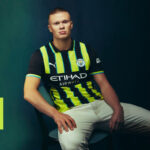 Young man modeling a Manchester City football jersey with neon-yellow and dark-blue vertical stripes, seated against a dark backdrop.