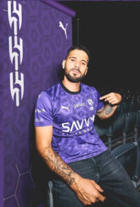 Man with a beard wearing a purple training jersey sits in stadium seats beside a purple banner with white logos; visible tattoos on his right arm.