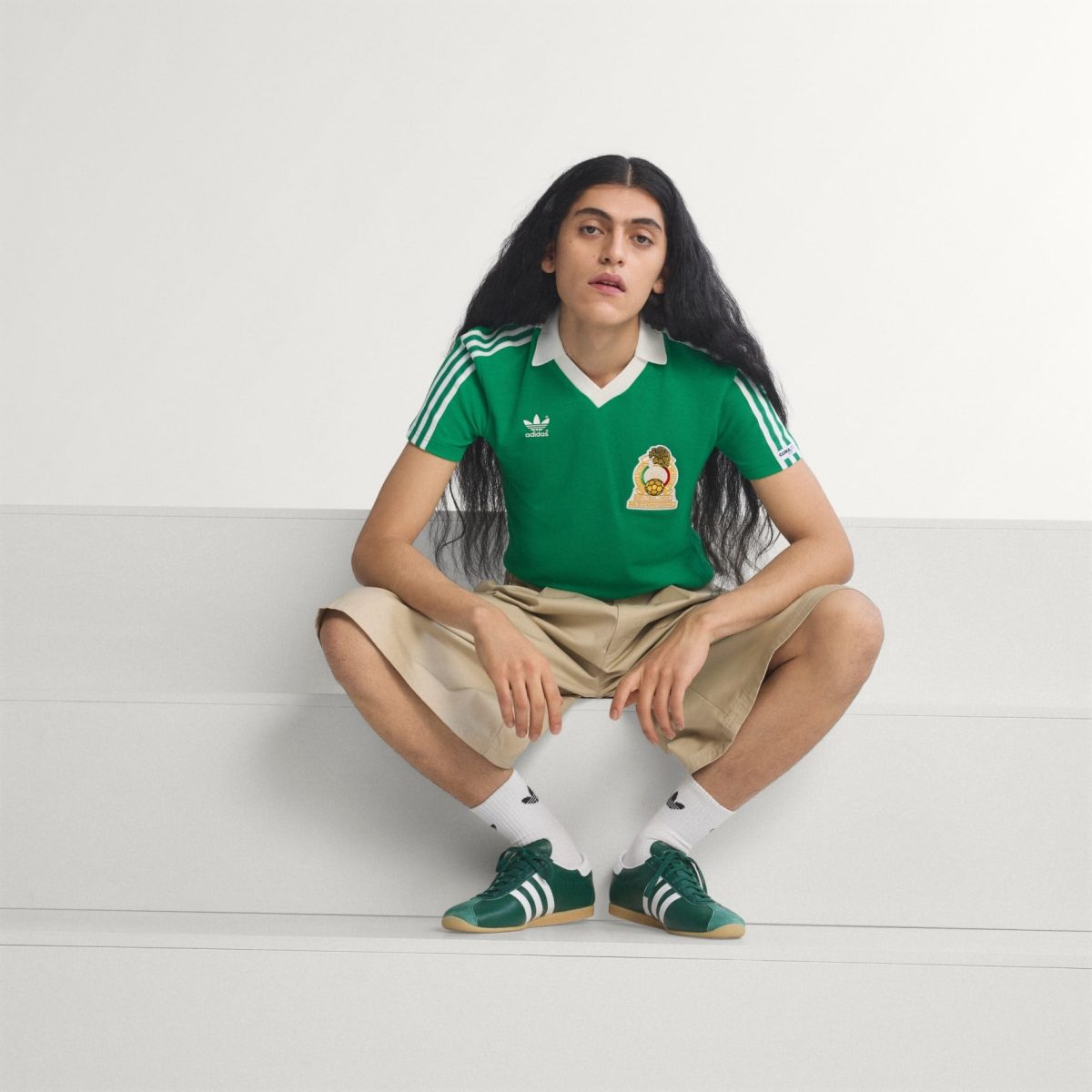 Woman with long black hair sits cross-legged on a white bench, wearing a green soccer jersey, beige shorts, white socks, and green sneakers.