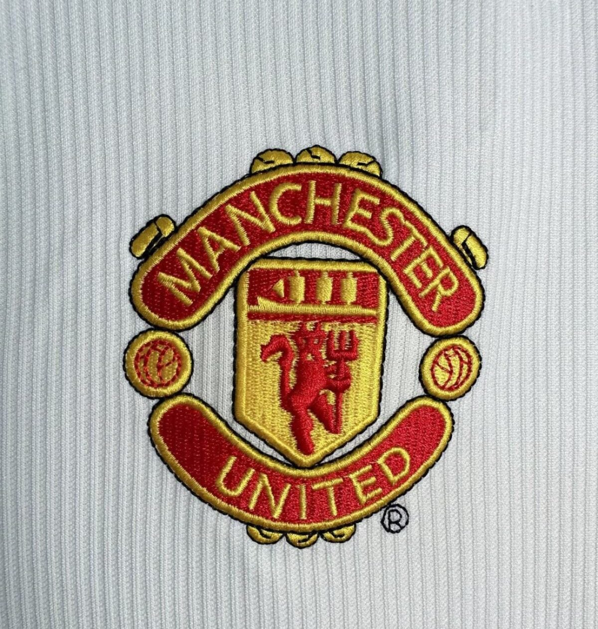 Embroidered Manchester United crest in red and gold on light gray ribbed fabric.