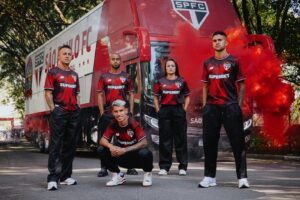 Five soccer players in red-and-black SPFC jerseys pose in front of a team bus, red smoke billowing nearby, outdoors.