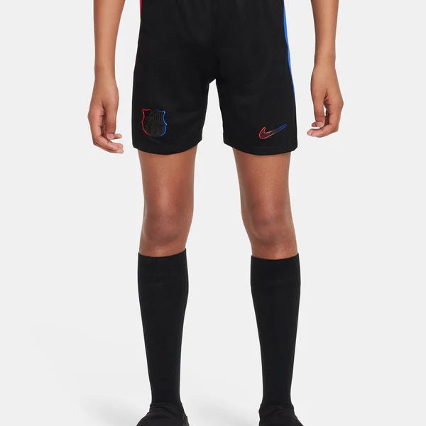 Black soccer shorts with red and blue side panels, small club crest on the left leg and a red Nike swoosh on the right, worn with black knee-high socks and cleats by a young player.