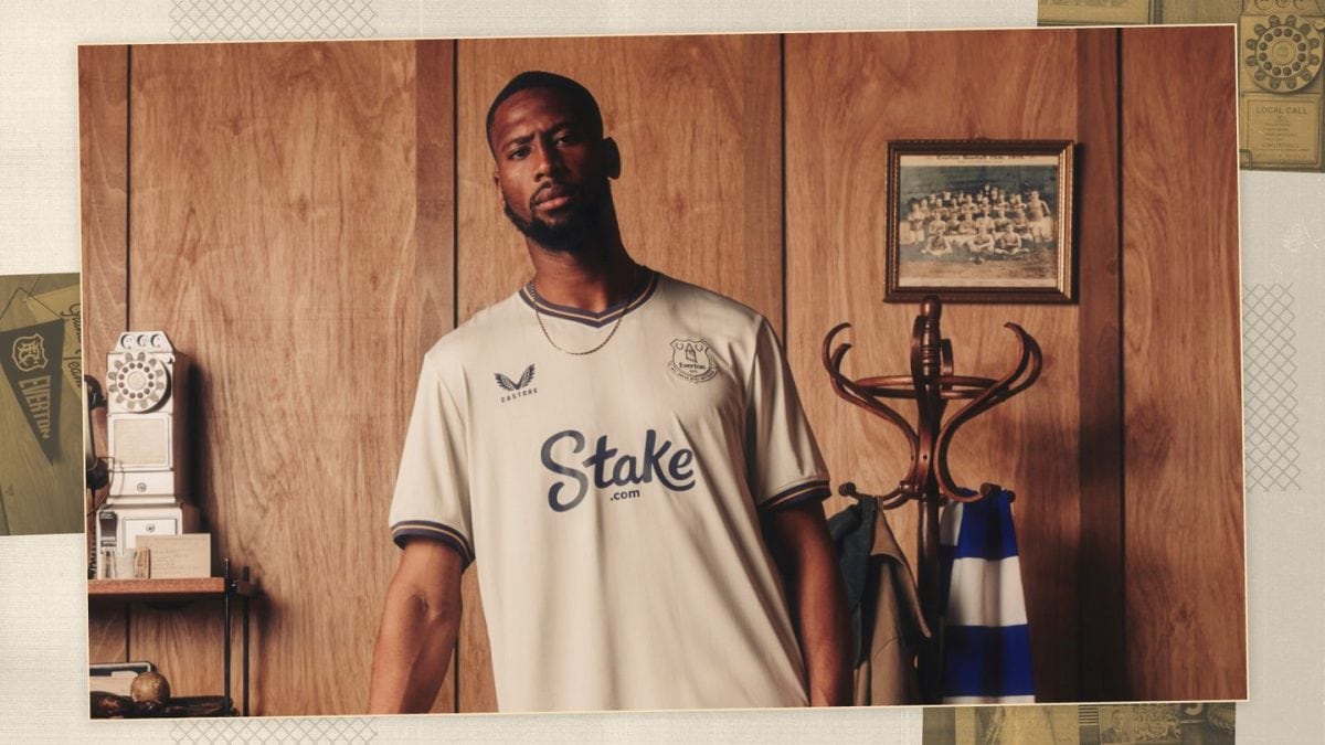 Man in a white Stake soccer jersey stands in a wood-paneled room with a coat rack and vintage phone.