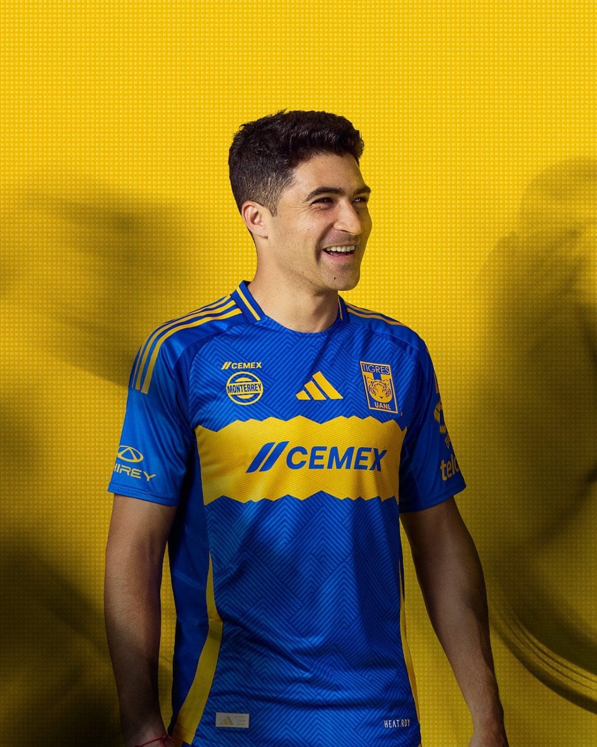 Portrait of a man in a blue and yellow Tigres UANL soccer jersey smiling against a bright yellow background with a patterned backdrop.