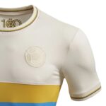 Close-up of a white soccer jersey showing an embossed crest on the chest, rainbow-trimmed collar and sleeve, with a yellow band and blue hem at the bottom