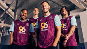 Four male soccer players pose in a locker room, wearing maroon jerseys with light blue sleeves and the 96.com logo on the chest, smiling at the camera.