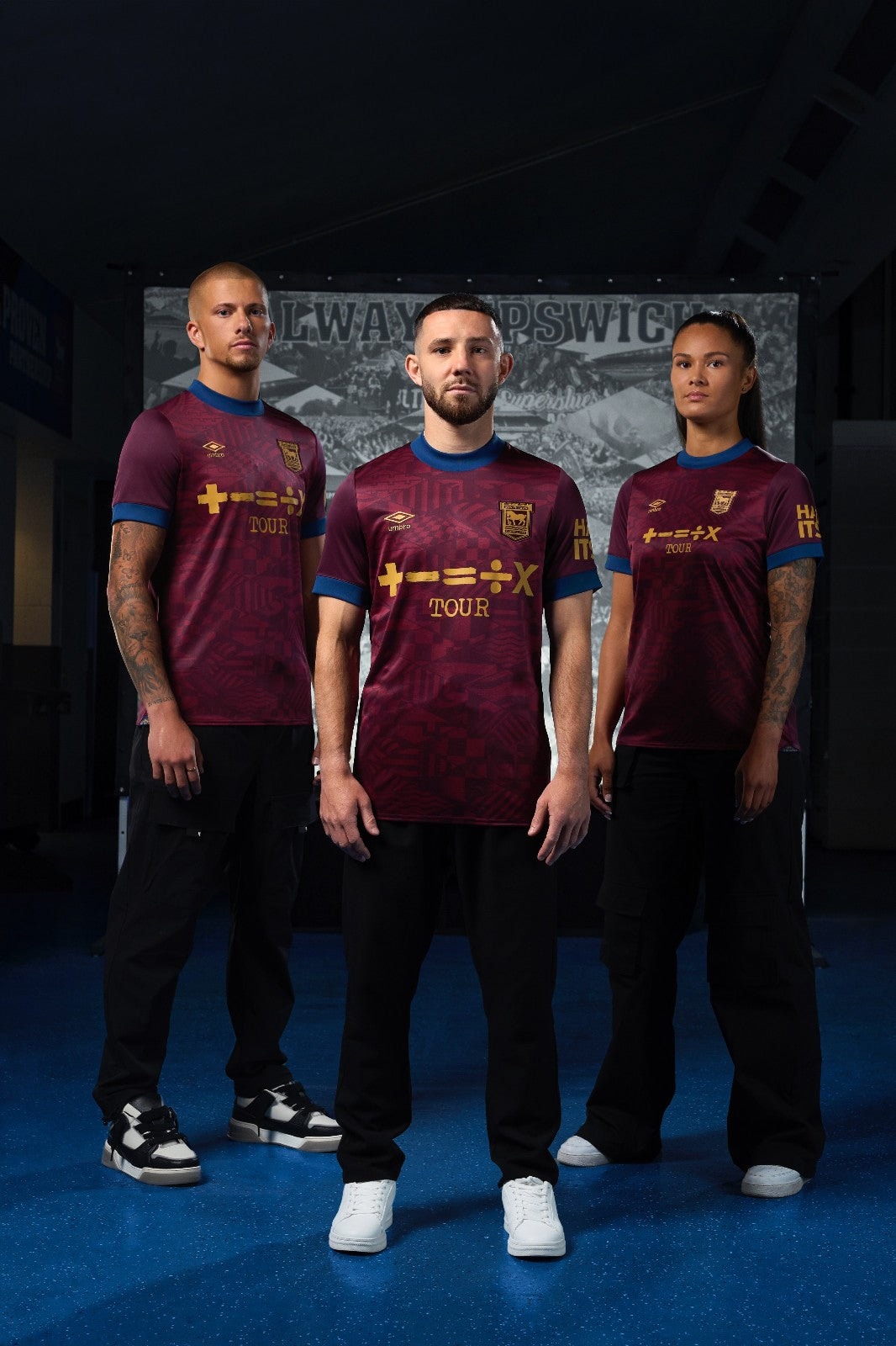FxIGZV3YgrjJSfW | UAE SPORT STORE Three athletes in maroon jerseys with blue trim stand in a dark tunnel, facing the camera.
