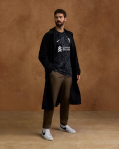 G0kFX12ov56xFK9_56b207b7-107a-4d1c-9836-628da60b11c0 | UAE SPORT STORE Man posing in a long black coat over a dark patterned athletic shirt, brown trousers, and white sneakers against a brown backdrop.