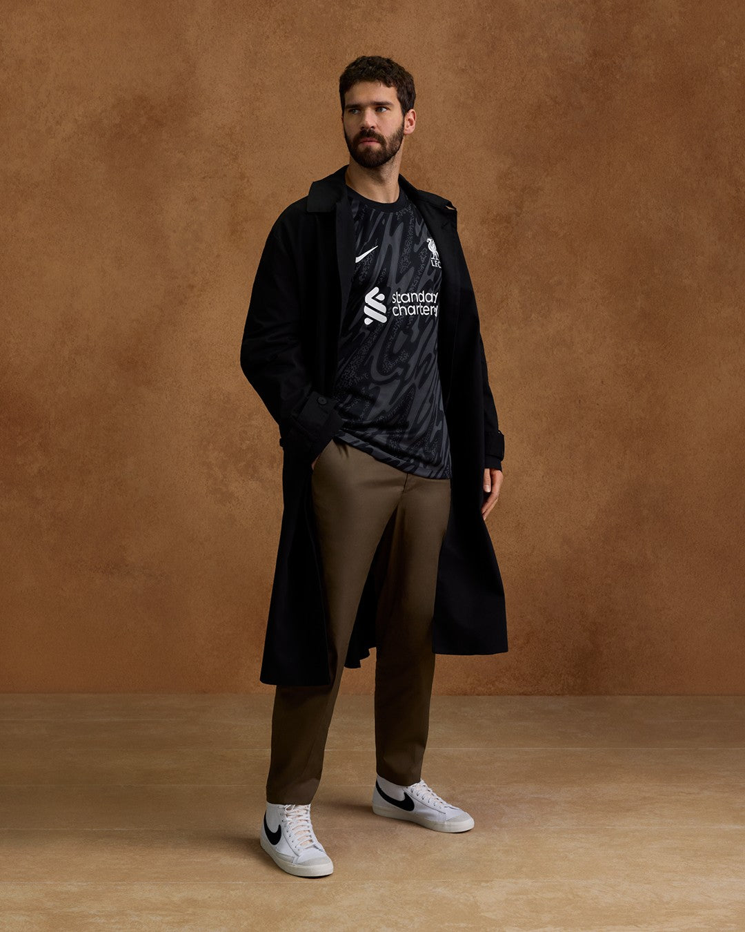 G0kFX12ov56xFK9_56b207b7-107a-4d1c-9836-628da60b11c0 | UAE SPORT STORE Man posing in a long black coat over a dark patterned athletic shirt, brown trousers, and white sneakers against a brown backdrop.