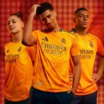 GAH9rdXBr6L1GBE | UAE SPORT STORE Three young athletes pose in orange Real Madrid jerseys with Emirates Fly Better logo against a red backdrop.