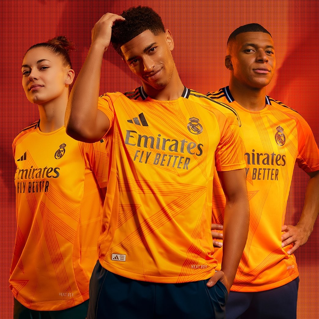 GAH9rdXBr6L1GBE | UAE SPORT STORE Three young athletes pose in orange Real Madrid jerseys with Emirates Fly Better logo against a red backdrop.