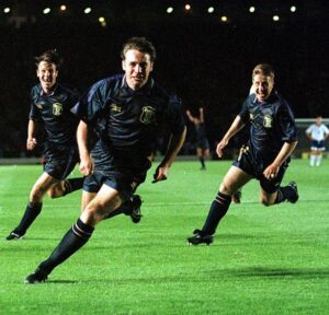 Three soccer players in dark uniforms sprint across a well-lit green field at night.