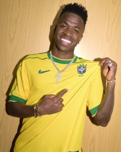 Smiling man in a Brazil soccer jersey points to a silver '7' pendant on his chain against a wooden background.