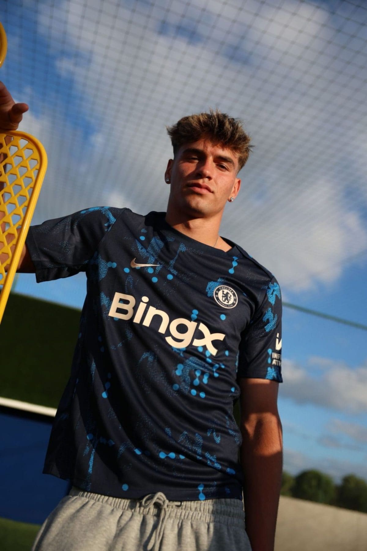 Young man in a Chelsea football jersey holds a yellow lacrosse stick, standing on a sunny field with a chain-link fence behind him.