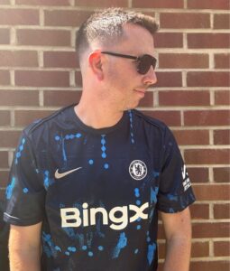 GS3EHLmXAAE86PU | UAE SPORT STORE Man wearing a dark Chelsea FC jersey with Sunglasses, standing against a brick wall, looking to the right.