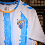 White Málaga CF football jersey with blue geometric patterns, orange collar trim, and club crest on the chest.