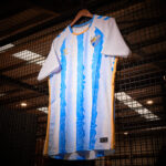 Sports jersey on a wooden hanger against a grid backdrop, featuring light blue and white vertical stripes with gold side panels and team crest.