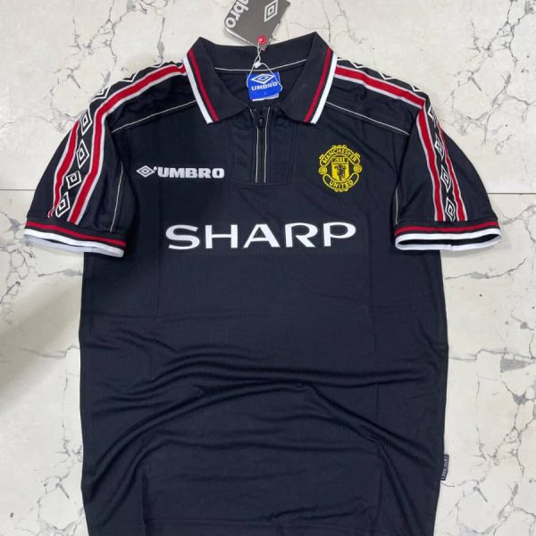 Dark Manchester United jersey on hanger featuring crest, Umbro logo, and 'SHARP' sponsor; navy with red-white trim.