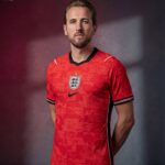 G_7O7WAX0AA31jf | UAE SPORT STORE Portrait of a man wearing a red England football jersey, facing the camera with a neutral expression against a dark gradient background