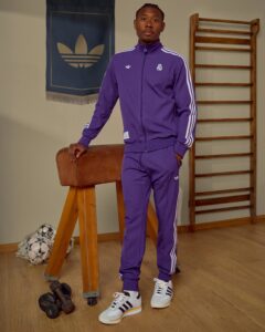 Young man in a purple Real Madrid tracksuit stands indoors by a wooden gym ladder, with soccer balls and dumbbells on the floor and an Adidas banner on the wall behind him.