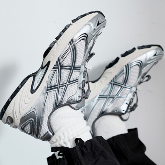 Giay-Asics-Gel-Kahana-TR-V4-Silver-White-1203A497-200-3-600x531-1 Two silver-gray running shoes with patterned soles, feet crossed in the air against a light background.