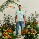 Glc8nhUWsAAEqyP_a5599c6e-e0c2-44df-b9ea-bdc7a4fa9c12 | UAE SPORT STORE Man in a light green Adidas T-shirt and blue jeans stands among vibrant orange and white flowers against a pale wall.