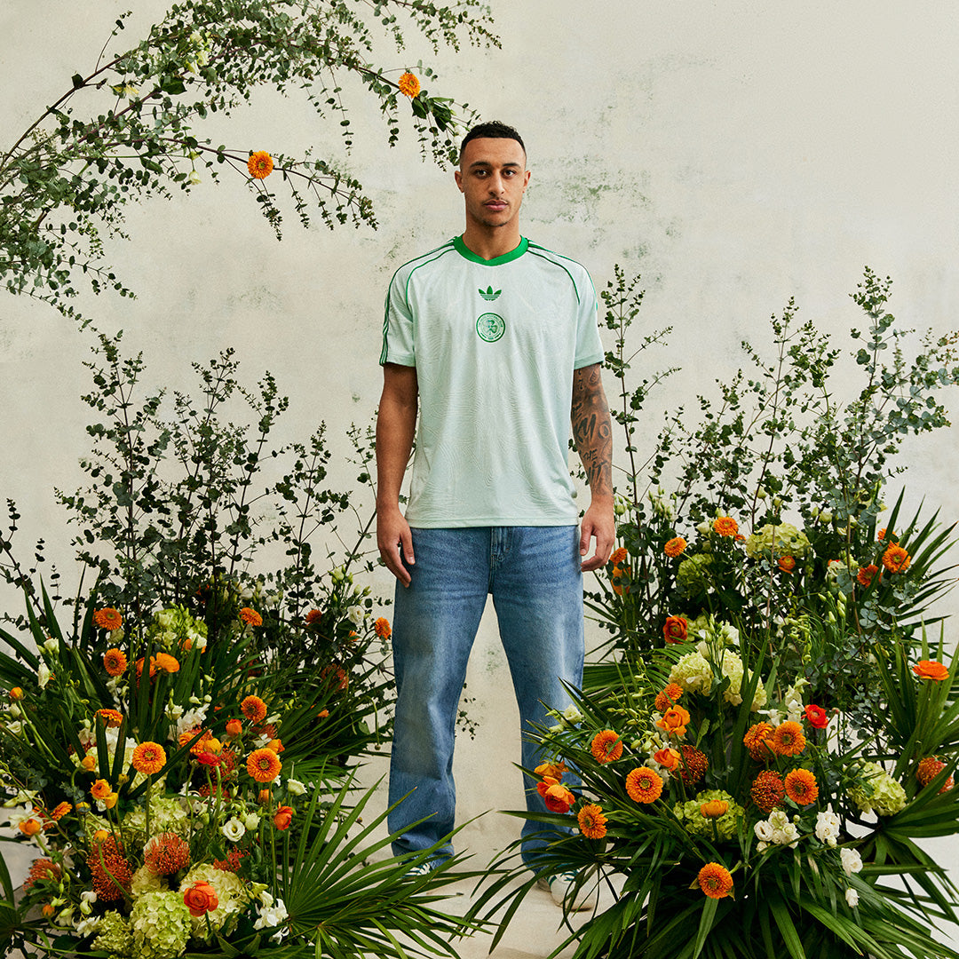 Glc8nhUWsAAEqyP_a5599c6e-e0c2-44df-b9ea-bdc7a4fa9c12 | UAE SPORT STORE Man in a light green Adidas T-shirt and blue jeans stands among vibrant orange and white flowers against a pale wall.