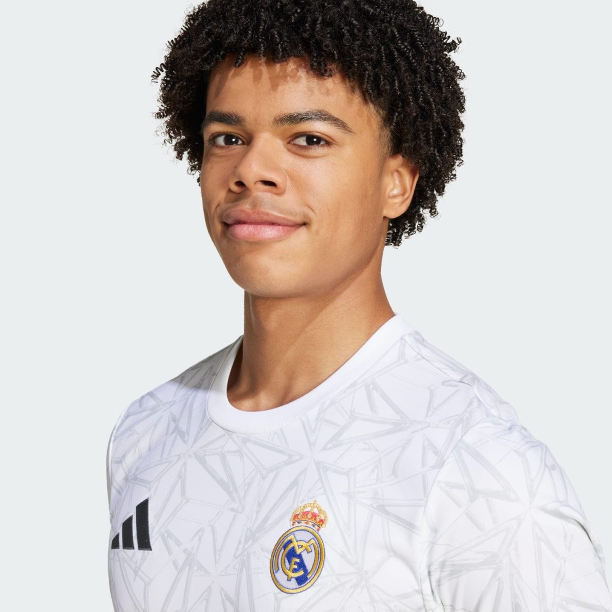 Young man wearing a white Real Madrid jersey with a geometric pattern, facing camera against a light gray background.