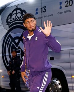 Footballer in a purple Real Madrid tracksuit waves as he exits a team bus with the club crest visible behind him.