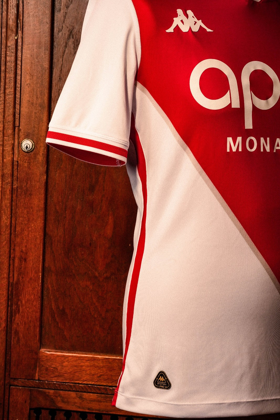 GvHAbflyHaShHSz | UAE SPORT STORE Close-up of a red and white sports jersey hanging against a wooden door, featuring the Kappa logo on the shoulder and partial text/logo on the front.