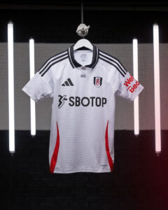 GvRDYQgOHJoZdGK | UAE SPORT STORE White Fulham FC jersey on a hanger with black triple stripes, crest, and SBOTOP sponsor against a backdrop of vertical neon lights.