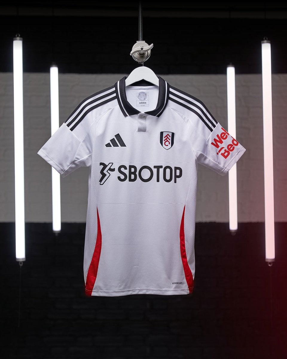 GvRDYQgOHJoZdGK | UAE SPORT STORE White Fulham FC jersey on a hanger with black triple stripes, crest, and SBOTOP sponsor against a backdrop of vertical neon lights.