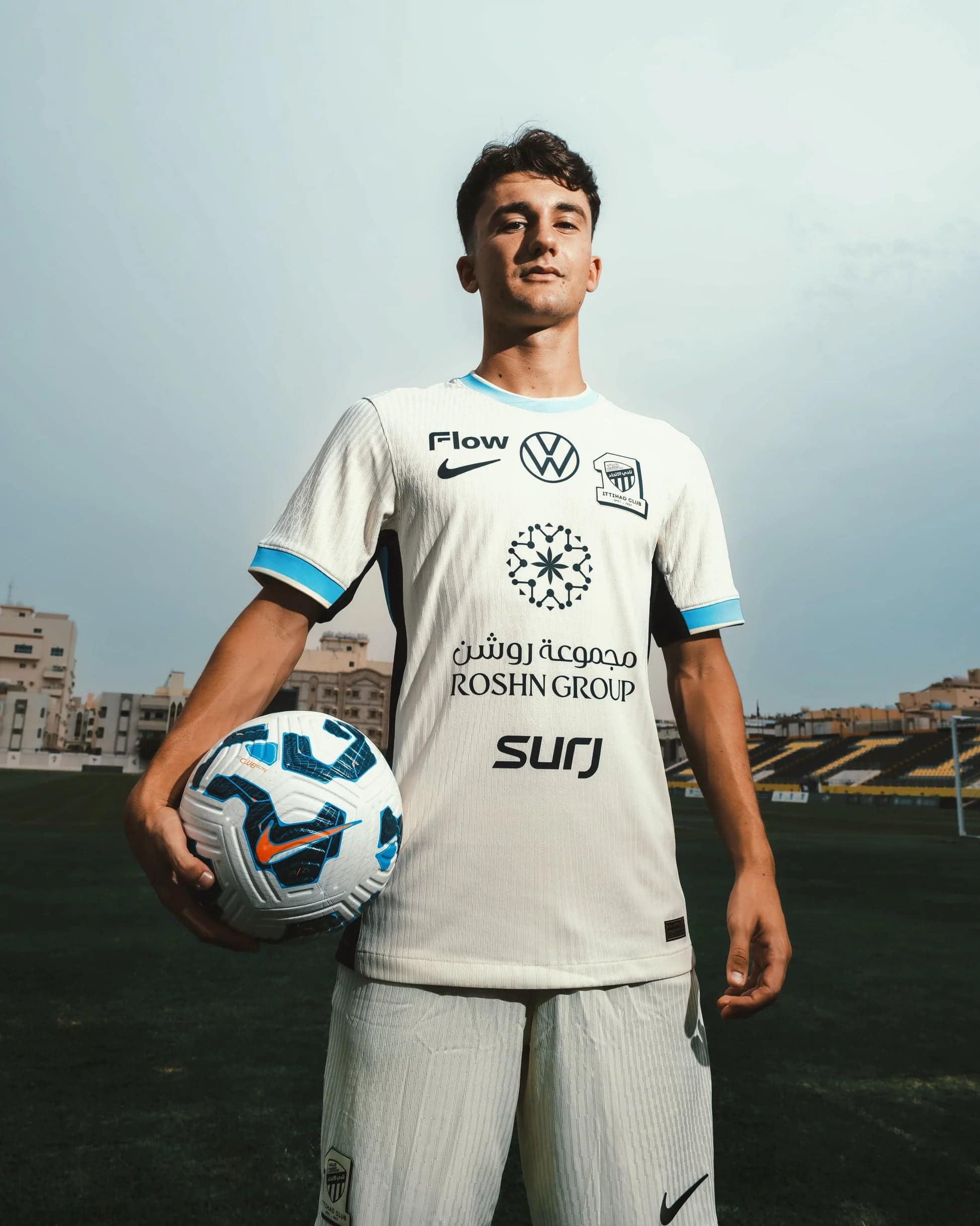 GygAIEwXkAA-nqO | UAE SPORT STORE Soccer player in a white jersey holding a blue and white soccer ball on a football field with stands in the background.