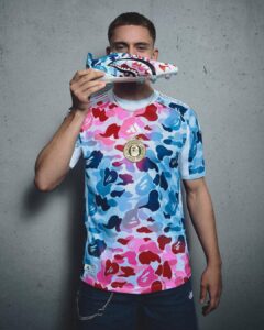 Young man in a multicolor camo Adidas jersey holds a matching sneaker up to his mouth against a gray wall.