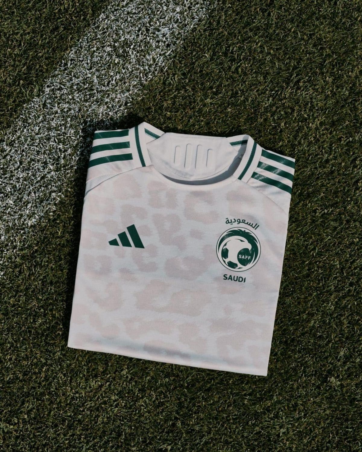 White Saudi football jersey folded on a grassy field, with SAFF logo and Arabic text visible on the chest area.