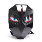 Black geometric mask with angular facets, glowing blue and red LED eyes, and a vertical LED strip on the forehead, placed on a white surface with straps visible underneath.
