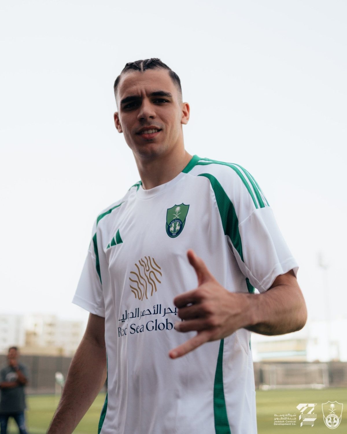 Smiling young man in a white jersey with green stripes giving a thumbs up on a sports field, crest on chest visible.